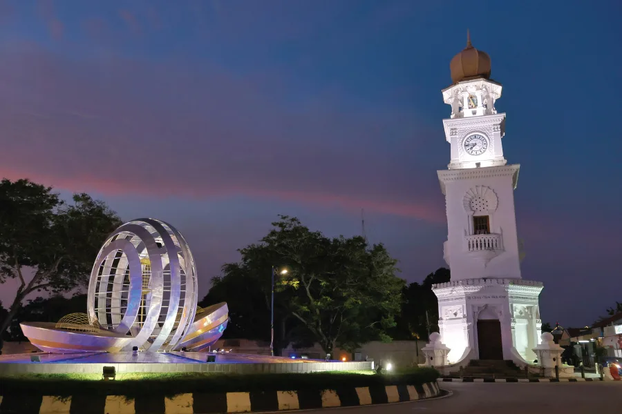 A clock tower and a sculpture in a roundabout which is illuminated with lights